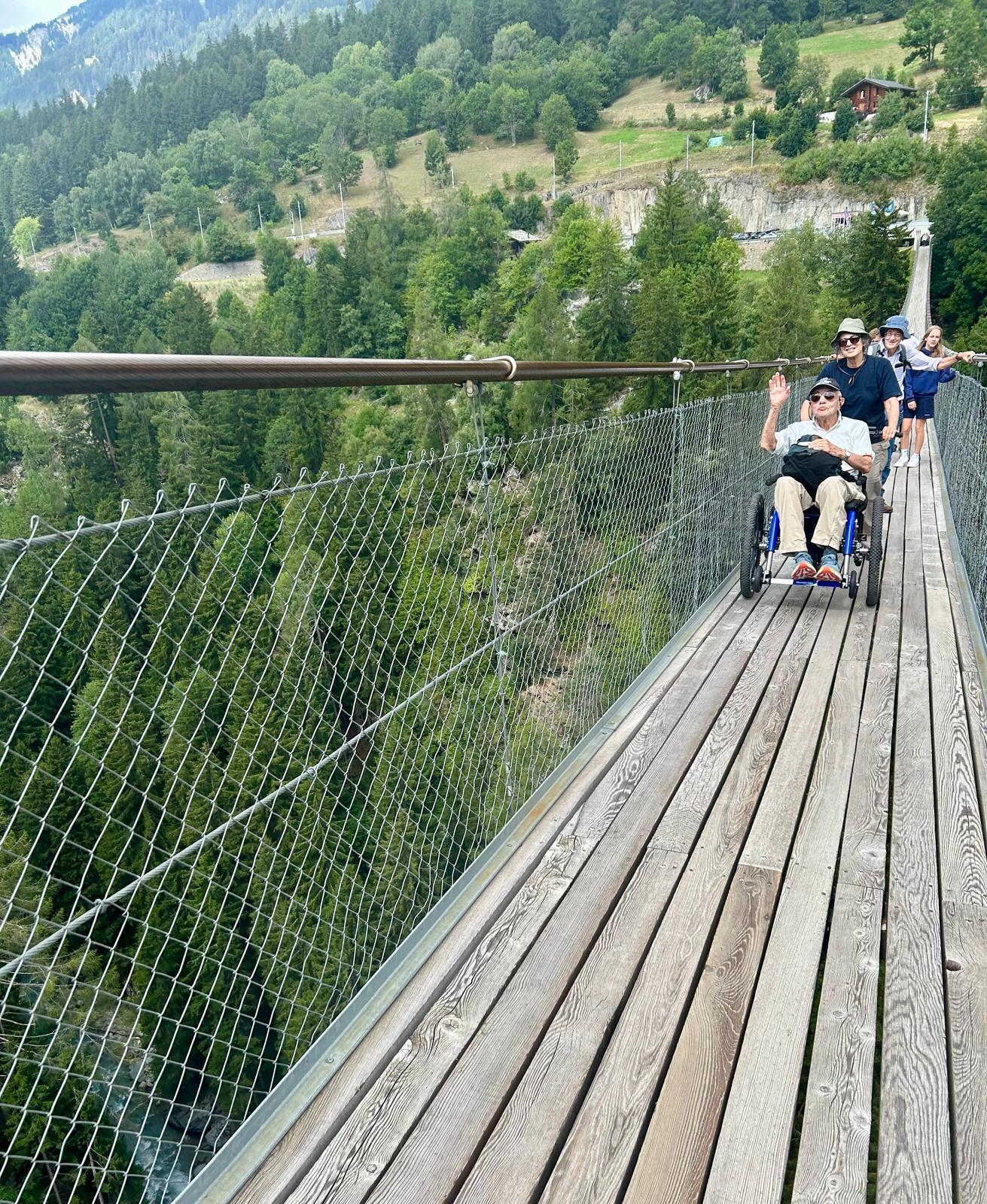 gentleman sat in an all terrain wheelchair being pushed along a long wooden bridge over a ravine