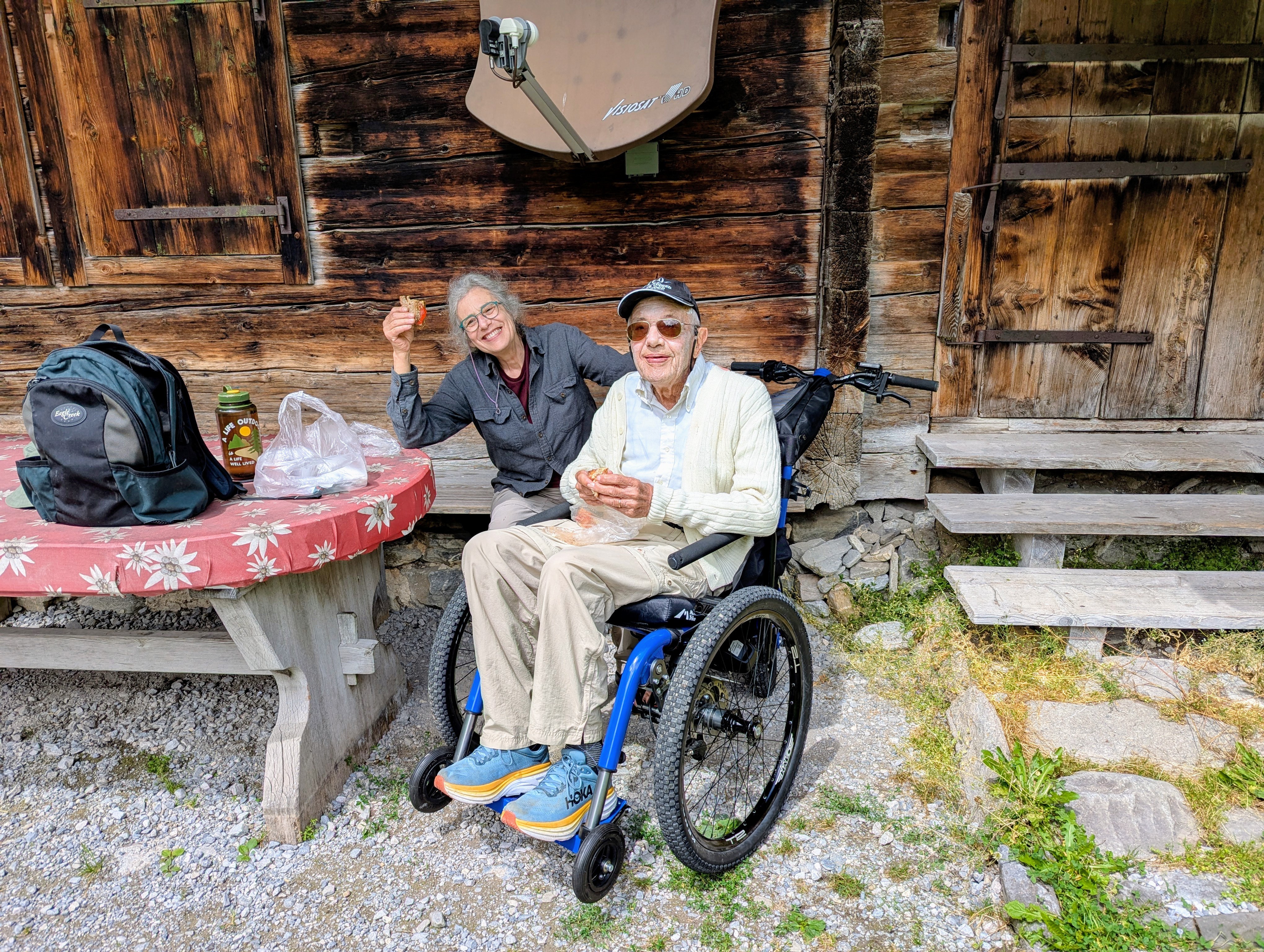 woman sat on bench eating a picnic with man sat in an all terrain wheelchair outside a wooden chalet