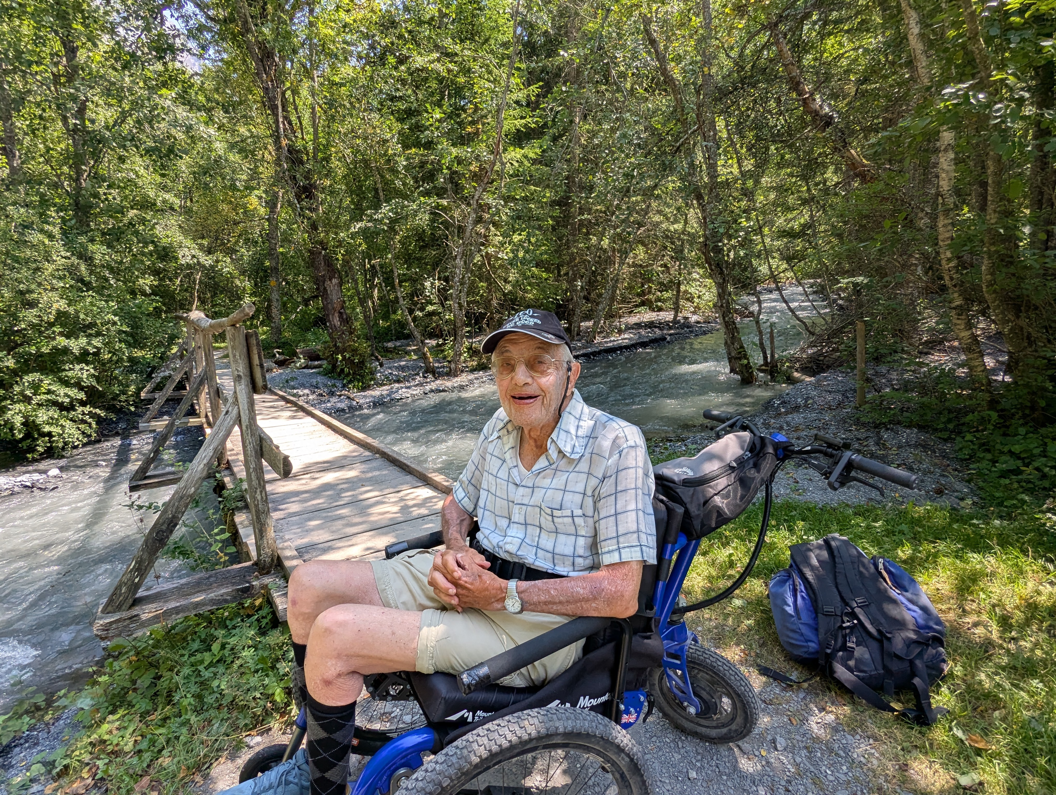 man sat in an all terrain wheelchair in the outdoors next to a river and wooden bridge