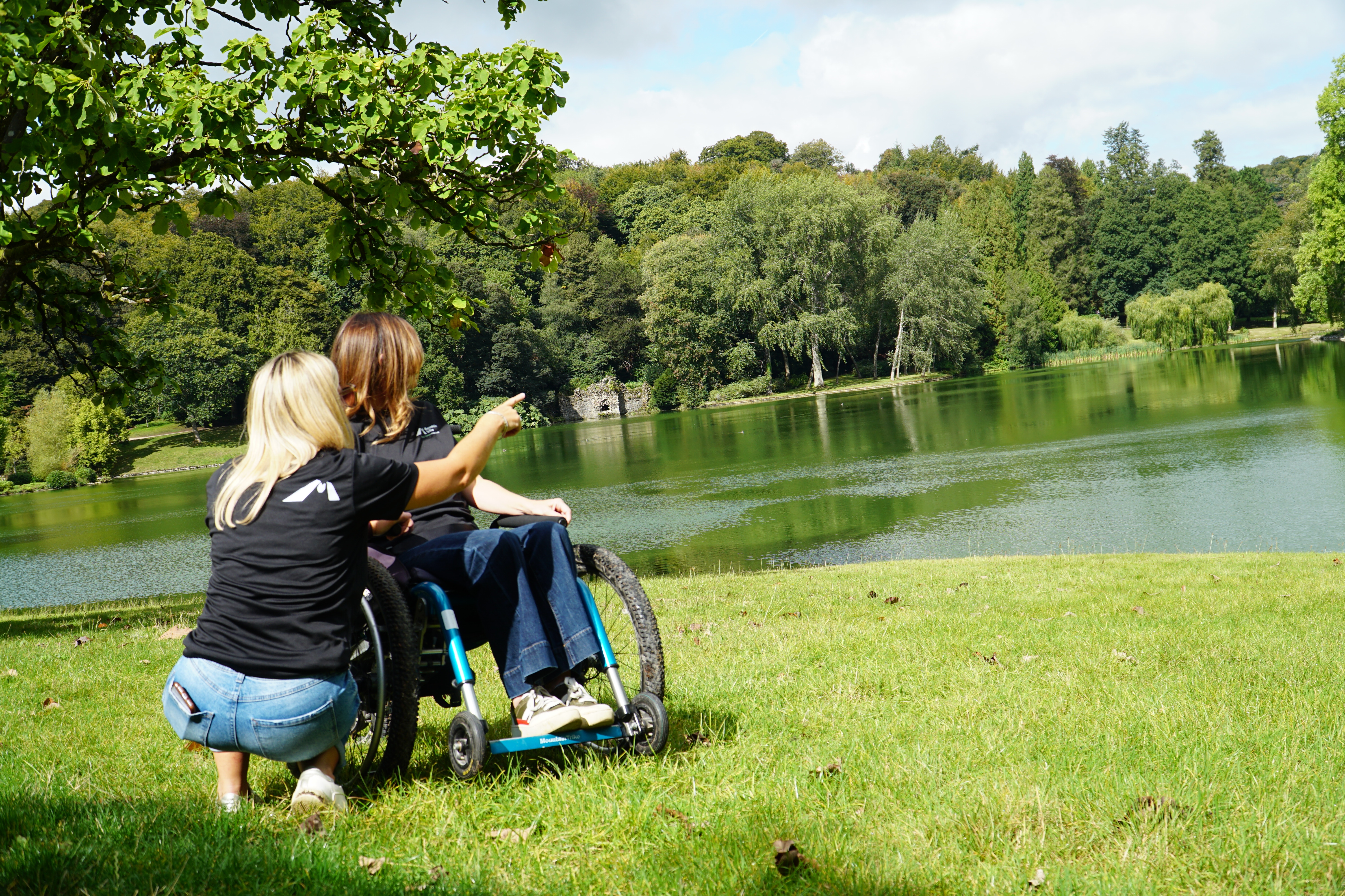 Two people looking out across a lake enjoying the views one person is crouching down pointing at the view, the other person is sat in an all terrain wheelchair looking at the same view