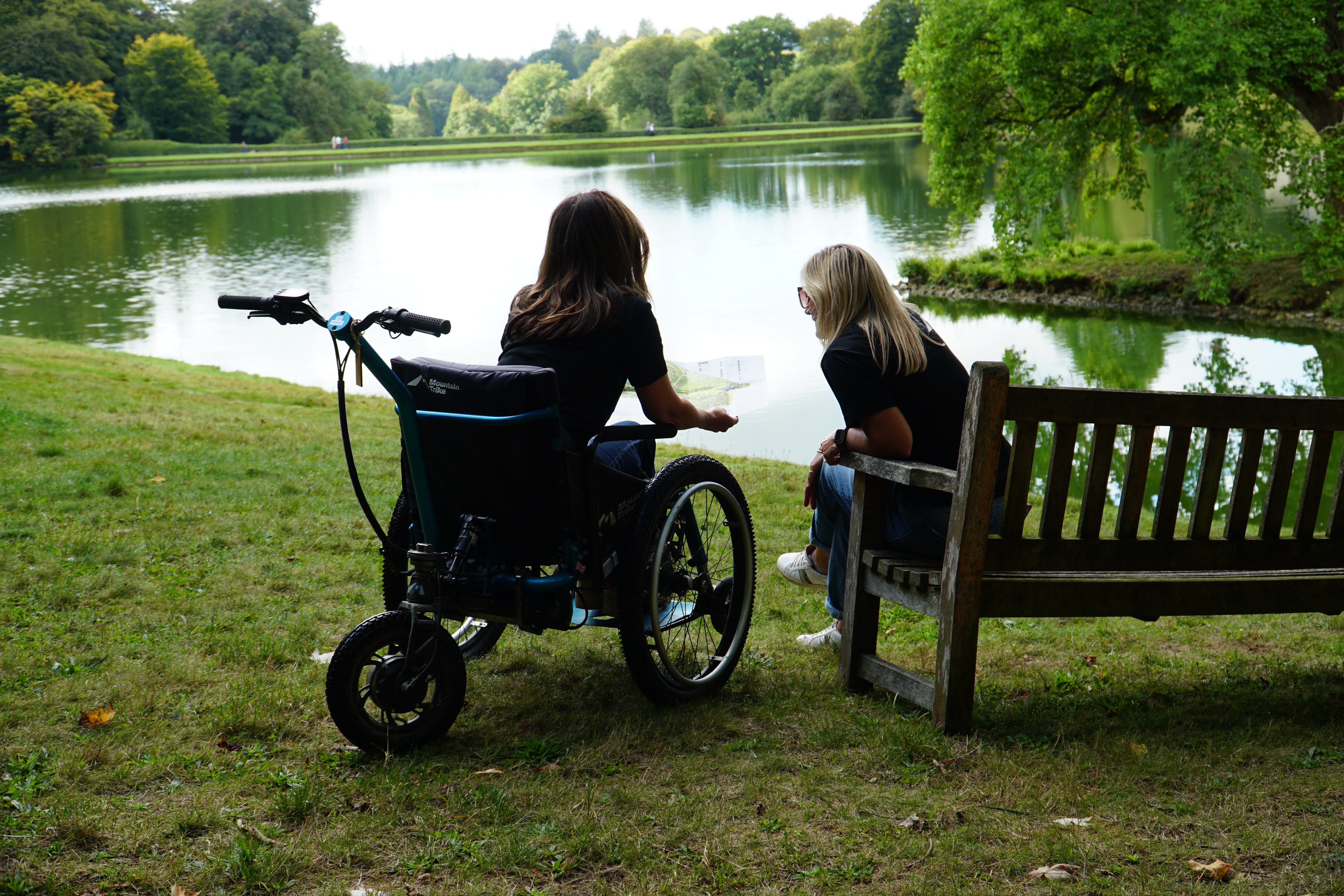 Two people in a parkland overlooking a lake looking at a map, one sat on a bench and the other in an all terrain wheelchair