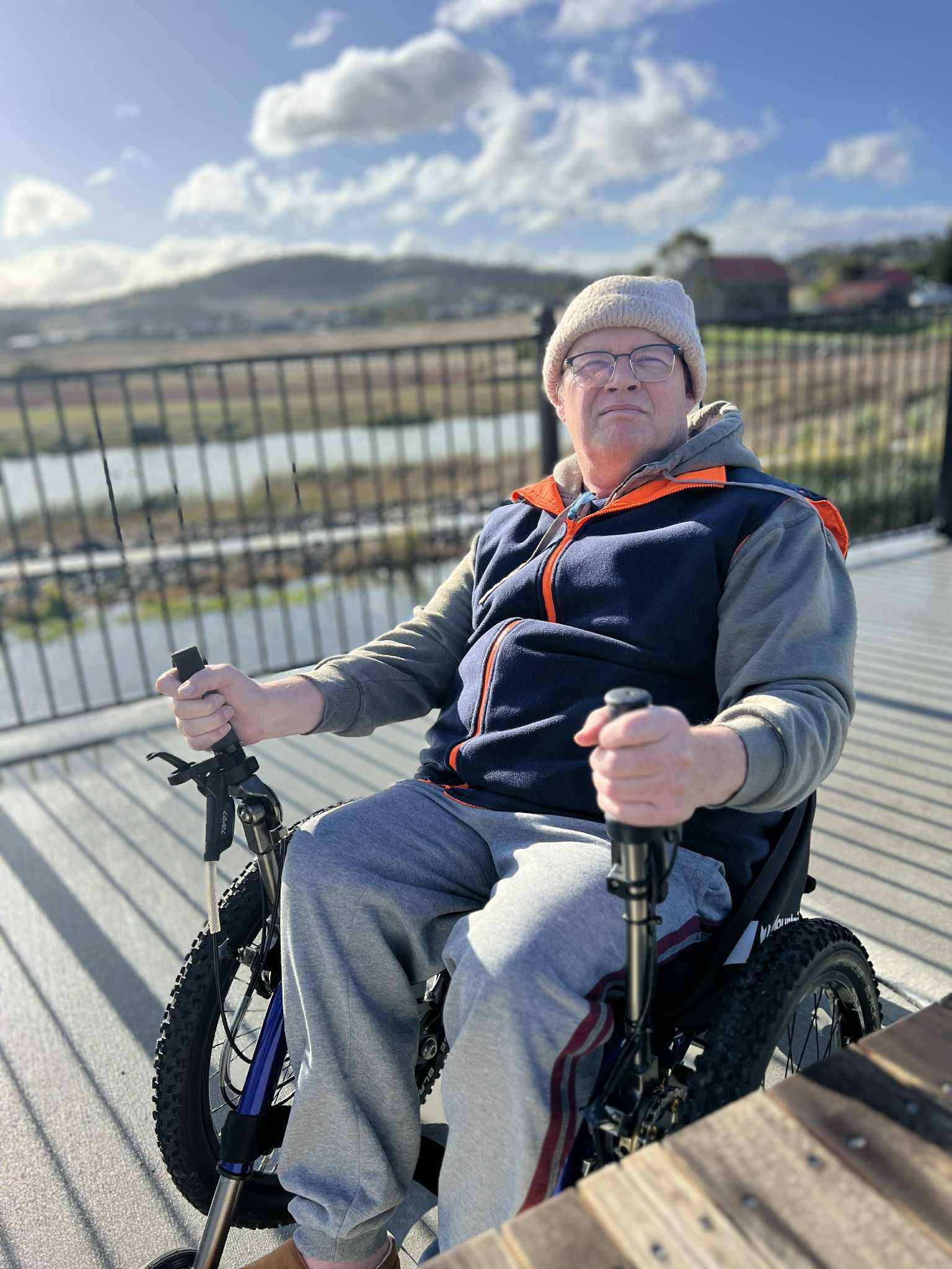 man sat in a mountain trike all terrain wheelchair on a deck with view of mountain in background